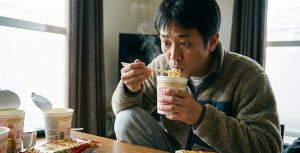 自宅のリビングルームで、フリースジャケットを着た日本人男性が、湯気の立つカップラーメンを箸で持ち上げ、目を見開いて恐る恐る口に運ぼうとしている様子。テーブルには他にも食べ終わったカップ麺や袋麺が散乱している。