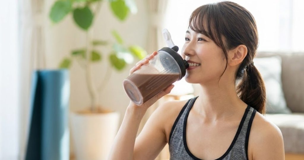 トレーニングウェアを着た若い日本人女性が、明るいリビングルームでシェイカーからチョコレート味のプロテインを飲んで笑顔を見せている様子。
