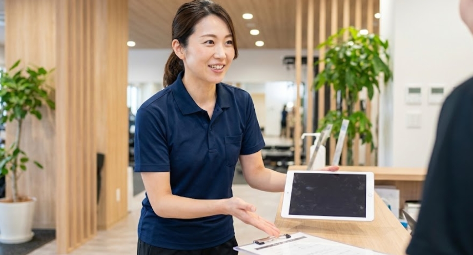 ジムの受付カウンターで、笑顔でタブレットを指し示しながら料金プランについて説明している日本人女性スタッフ。
