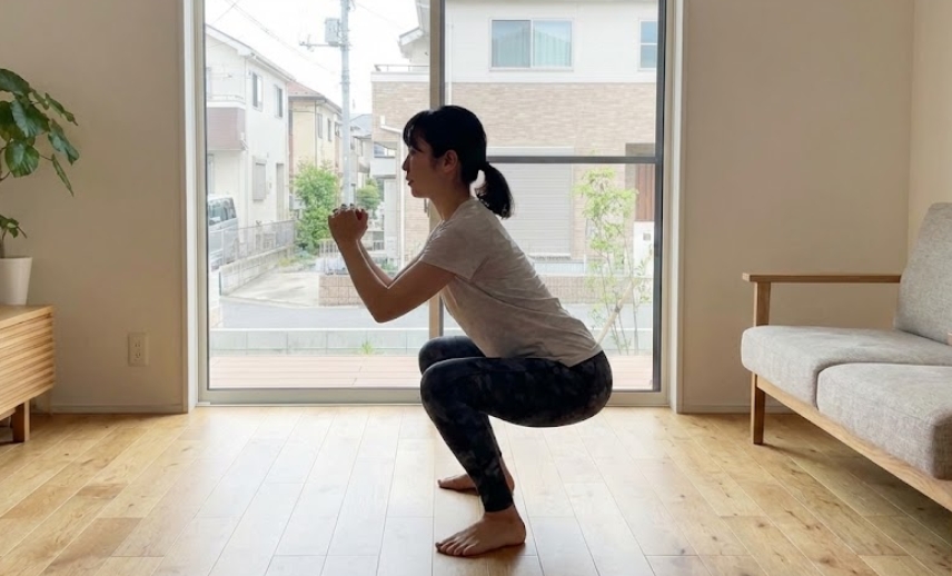 明るいリビングルームで、正しいフォームで深く自重スクワットを行っている日本人女性の横顔