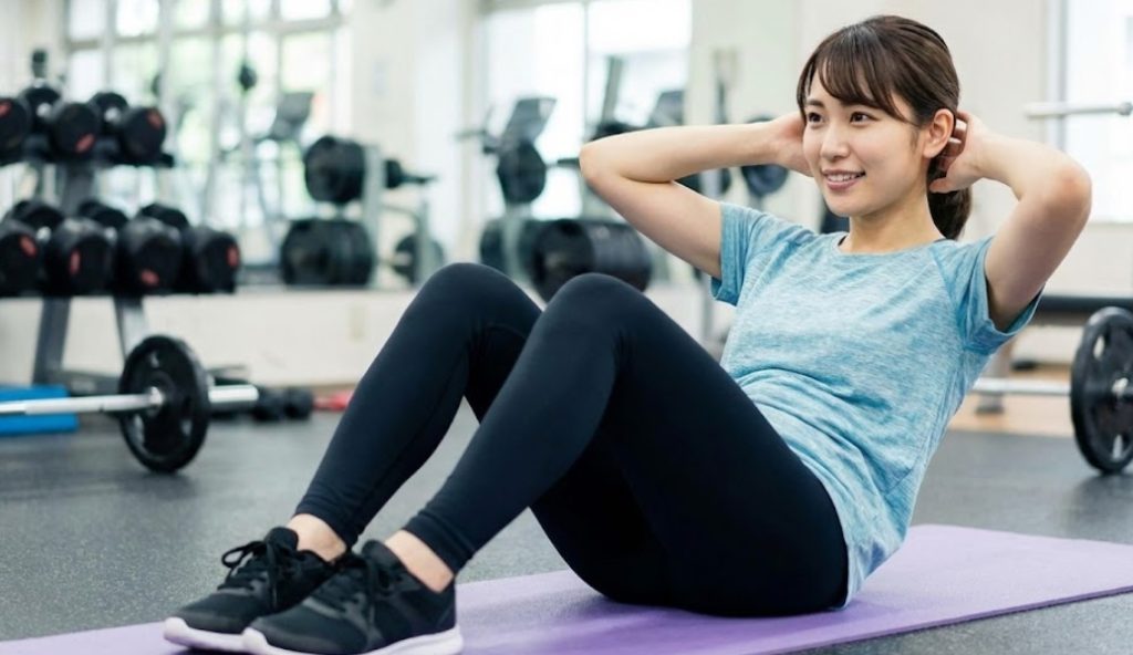 明るいフィットネスジムで、笑顔でクランチ（腹筋運動）を行っている日本人女性の様子。背景にはダンベルやトレーニングマシンが並んでいる