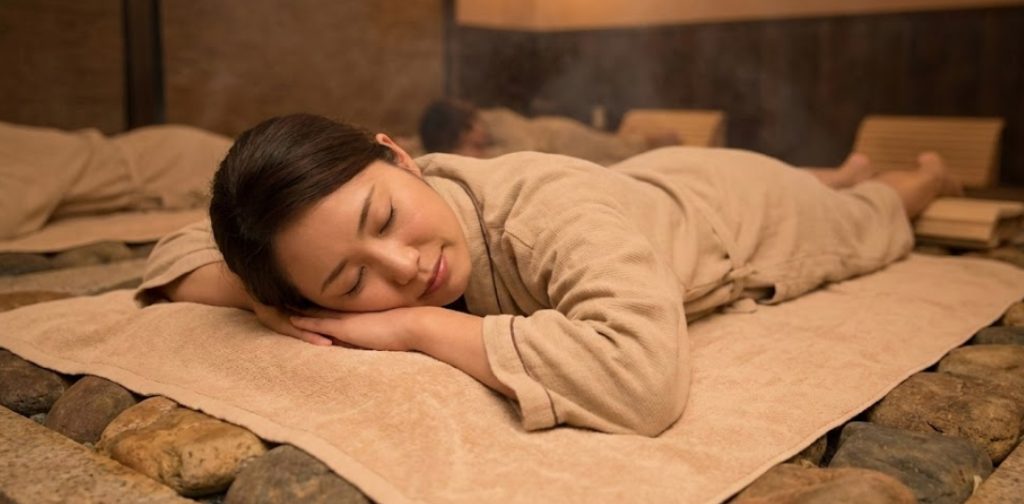 岩盤浴着を着て、温かい石の上に敷いたタオルの上でうつ伏せになり、目を閉じて気持ちよさそうにリラックスしている日本人女性の様子。