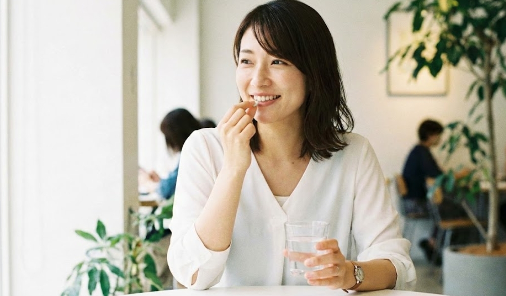 明るい自然光が入るカフェで、白いブラウスを着た笑顔の日本人女性が、コップの水と一緒に口臭ケアのためのサプリメント錠剤を口に運んでいる様子。