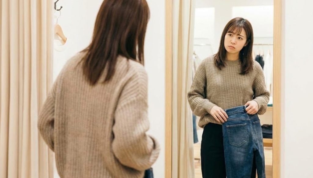 試着室の鏡の前でジーンズを腰に当て、ヒップの大きさを気にして不安そうな表情で悩んでいる若い日本人女性