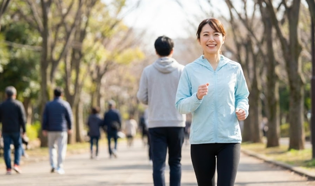 晴れた日の公園で、青いジャケットを着た若い日本人女性が笑顔でジョギングをしている様子。