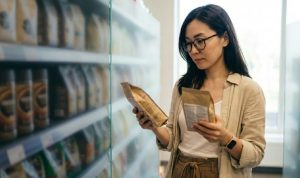 スーパーマーケットのコーヒー売り場で、メガネをかけたアジア系の女性が、左右の手に持った2種類のコーヒー豆の袋の裏面ラベルを真剣な表情で見比べている様子。