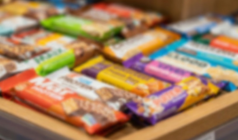 木製の棚に、オレンジ、紫、緑、黄色など、色鮮やかで多様なパッケージの海外製プロテインバーが並んでいる様子が、全体的にぼかして写されている。ブランド名やテキストは一切判読できない。