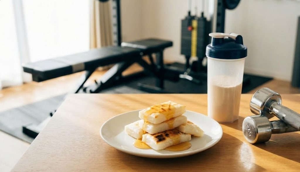 自然光が差し込むホームジムのテーブルに、はちみつがかけられた焼き餅の皿、プロテインシェイカー、ダンベルが置かれている。背景にはトレーニングベンチが見える。