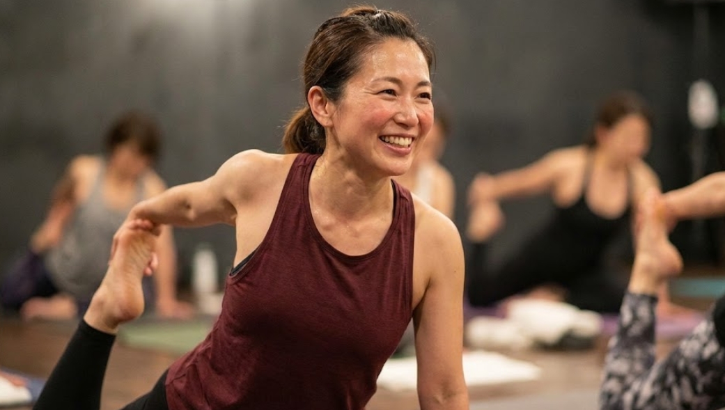 ホットヨガスタジオで笑顔でポーズをとる40代女性。汗をかきながら楽しそうにヨガに取り組んでいる。