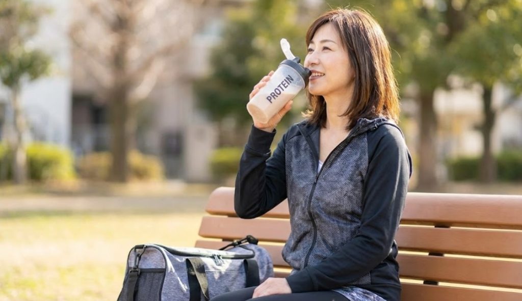 晴れた日の公園のベンチで、運動後に笑顔で「PROTEIN」と書かれたシェイカーからプロテインを飲んでいる、スポーツウェア姿の40代日本人女性。横にはジムバッグが置かれている。