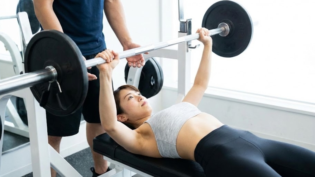 明るいジムで、後ろに男性の補助者（スポッター）をつけ、真剣な表情でバーベルベンチプレスを持ち上げている日本人女性