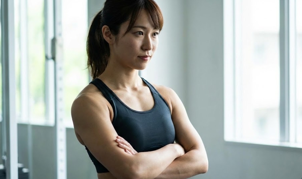 ジムの窓辺で黒いスポーツブラを着用し、腕を組んで立つ日本人女性。大胸筋のトレーニングによって引き締まったデコルテラインと、背筋が伸びた美しい姿勢が強調されており、自信に満ちた表情をしている。
