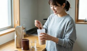 自然光が差し込む家のキッチンで、グレーのスウェットを着た日本人女性が、スプーンを使ってプロテインシェイカーにココアパウダーを入れている様子。