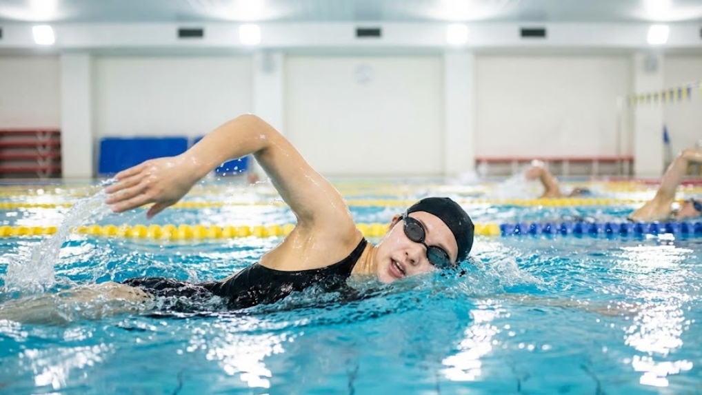 黒い競泳水着とスイムキャップ、ゴーグルを着用した若い日本人女性が、明るい照明の室内プールで水しぶきをあげながらクロールで泳ぎ、息継ぎをしている様子。背景のコースでは他のスイマーたちも練習している。