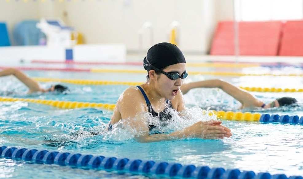 明るい室内プールで、黒いスイムキャップとゴーグルを着用した日本人女性が、水しぶきをあげながら平泳ぎで息継ぎをしている様子。背景のコースでは他の泳者も練習している。