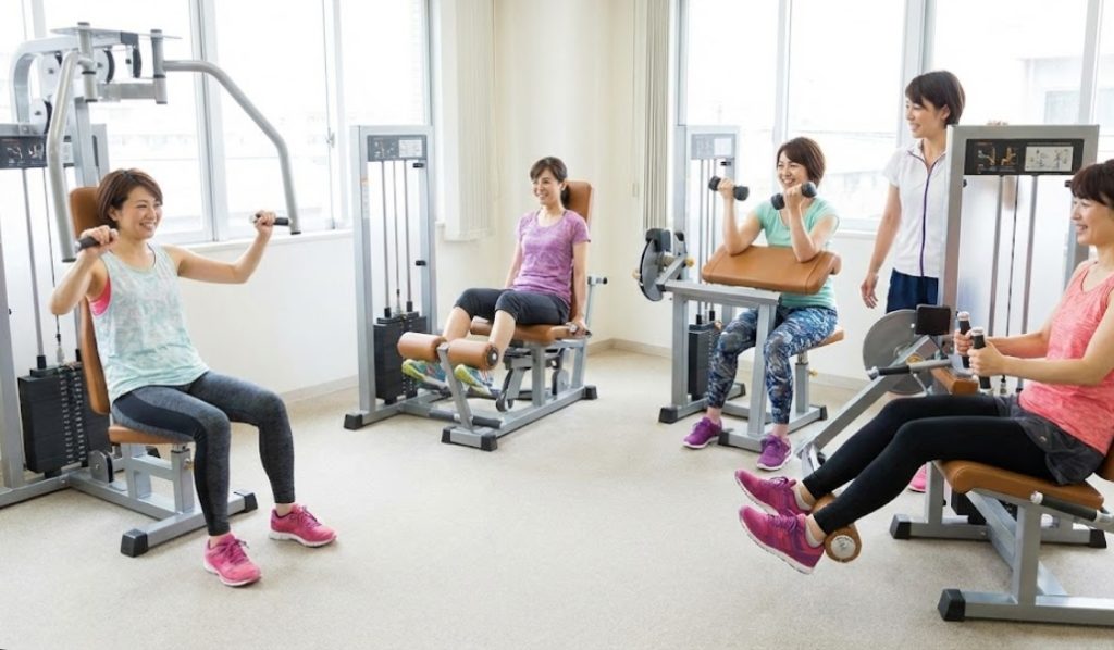 明るいフィットネスルームで、円形に配置された様々なウェイトマシンを使い、笑顔でサーキットトレーニングを行っている40代の日本人女性グループ。
