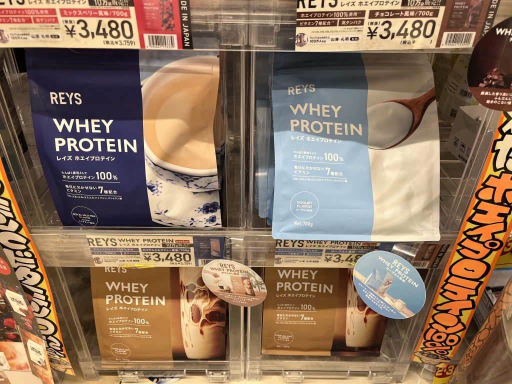 ドン・キホーテの棚に陳列されたREYSホエイプロテイン。ロイヤルミルクティー風味、ヨーグルト風味、カフェオレ風味が並び、価格は3,480円（税込3,759円）と表示されている。