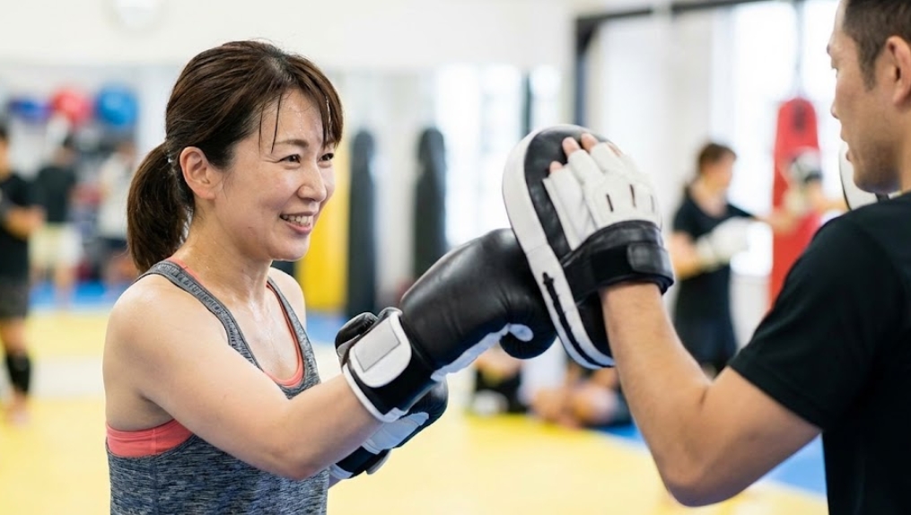 明るい格闘技ジムで、ボクシンググローブを着けた40代くらいの日本人女性が、トレーナーが持つミットに向かって笑顔でパンチを打っている。初心者が楽しそうにトレーニングしている様子。