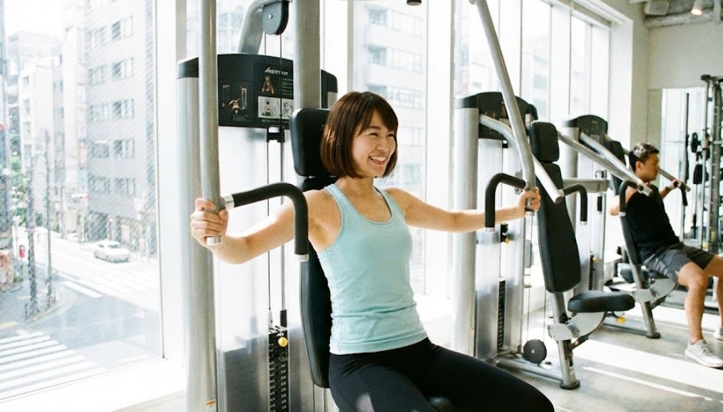 大きな窓から光が差し込む明るいジムで、水色のタンクトップを着た日本人女性が、満面の笑みを浮かべながらチェストプレスのマシンを使ってトレーニングをしている様子。