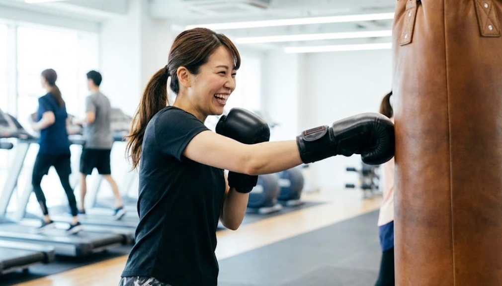 明るいフィットネスジムで、トレーニングウェアを着た日本人女性が、笑顔で楽しそうにサンドバッグにパンチを打ち込んでいる様子。フィットネス目的のキックボクシングトレーニング中。