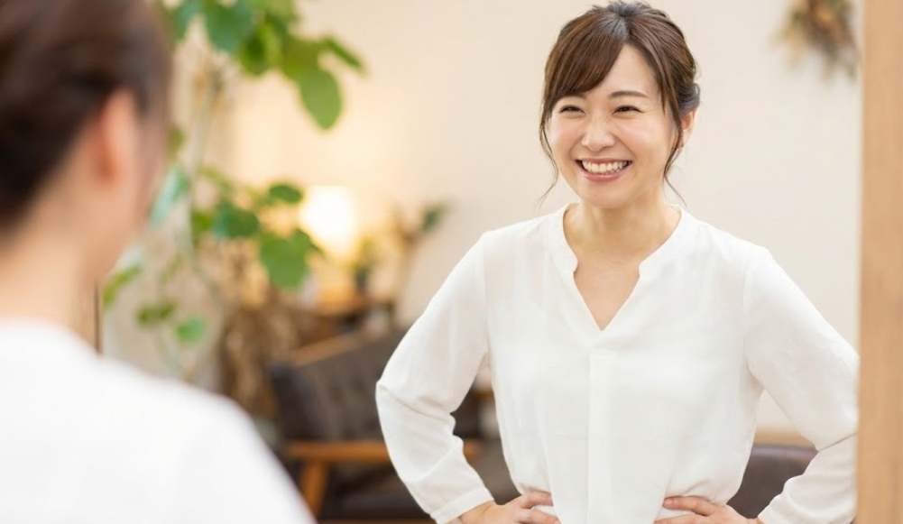 白いブラウスを着た30代前後の日本人女性が、鏡に映った自分の姿を見て満面の笑みを浮かべ、腰に手を当てて自信に満ちたポーズをとっている様子。背景は温かい雰囲気の部屋で、観葉植物やソファがぼけて見える。