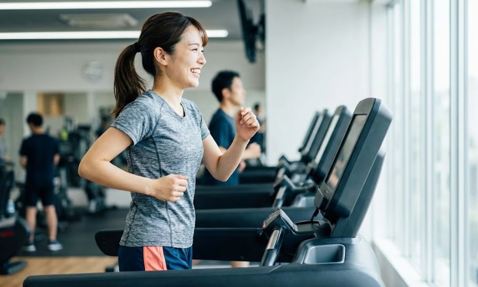 明るいフィットネスジムの窓際で、スポーツウェアを着た日本人女性が笑顔でルームランナー（トレッドミル）を使って走っている様子。