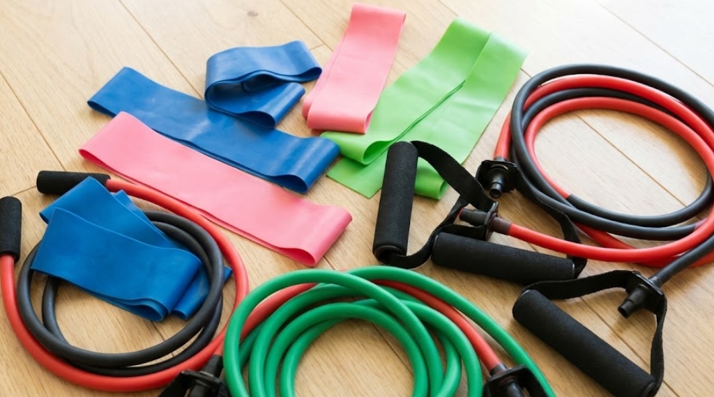 明るいフローリングの床に広げて置かれた、様々な種類と色のトレーニングチューブとレジスタンスバンドの俯瞰写真。青、ピンク、緑の平たいゴムバンド（セラバンドタイプ）と、持ち手（ハンドル）が付いた赤、黒、緑のチューブタイプが混在している。