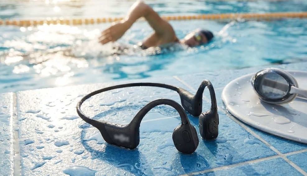 プールサイドの濡れた青いタイルの上に置かれた、黒いネックバンド型の防水骨伝導イヤホン。本体には水滴が付着しており、ロゴ部分はぼかされている。隣には白いプレートに乗ったスイミングゴーグルがあり、背景のプールでは人が泳いでいる。