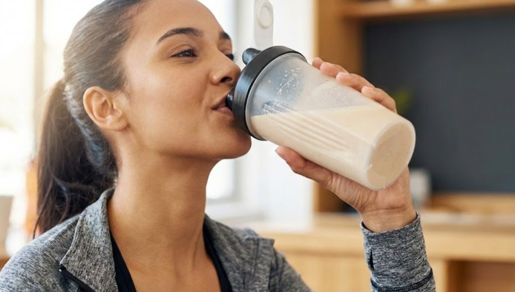 トレーニング後、明るいキッチンでシェイカーボトルからプロテインシェイクを飲むフィットネスウェアを着た女性の横顔。