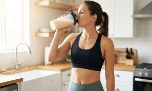 トレーニングウェアを着た女性が、明るい日差しの入るキッチンでシェイカーボトルからプロテインシェイクを飲んでいる様子。