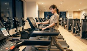 24時間ジム（夜）で、ワイヤレスイヤホンを装着し、自信を持って笑顔でトレッドミルを操作する、快適なフィットネスウェアを着た日本人の初心者女性。主体性と自分なりの通い方を見つけた様子を表現している。