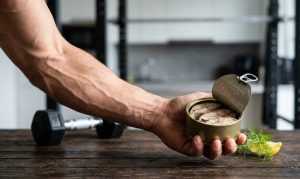 筋肉質で血管が浮き出たアスリートの腕が、暗い木製のテーブルの上に置かれた開いたサバ缶を持っている。缶の中には調理された魚が詰まっている。テーブルの左側にはシルバーのダンベルが1つあり、右側には新鮮なディルの小枝とレモンのスライスが添えられている。背景にはジムのパワーラックなどの設備がぼんやりと見え、トレーニングと栄養摂取の重要性を伝えている。