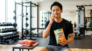 ジムで普通のポテトチップス（横のテーブル）ではなく、プロテインチップス（手に持つ袋）を笑顔で食べる日本人男性