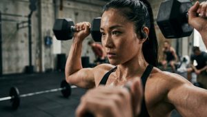 汗をかいて真剣な表情でダンベルプレスをする女性アスリート。耳にはしっかりと固定されたワイヤレススポーツイヤホンが装着されており、ジムの騒音をカットして集中している様子。