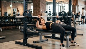 モダンなジムのフラットベンチで、リストラップと適切なフォームを保ちながら、バーベル（40kg目標）をベンチプレスする笑顔の日本人女性。自信と達成感を表している。