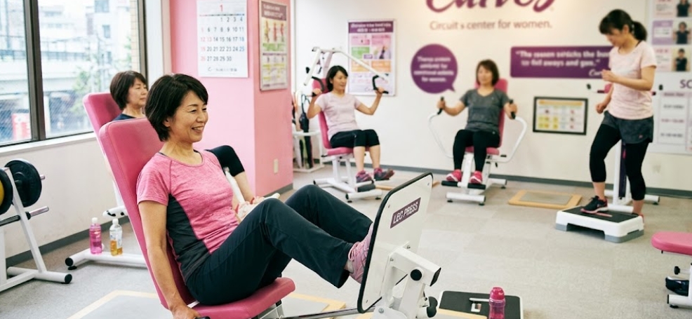 女性専用ジム「Curves」の明るい室内で、4人の女性が笑顔で異なるトレーニングマシンを使い、サーキットトレーニングを行っている。前景の女性はピンク色のTシャツを着てレッグプレスをしており、奥にはチェストプレスマシンやステップボードを使用する女性がいる。壁には「Curves」のロゴやカレンダー、スケジュール表などが貼られている。