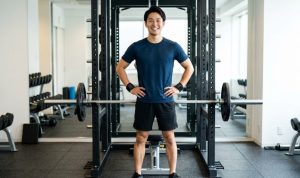 近代的で清潔なジムで、BIG3トレーニング（スクワット、ベンチプレス、デッドリフト）を終えた直後の、笑顔の日本人男性。両手を腰に当て、リストラップを着用。背景にはパワーラックとバーベルが見える。